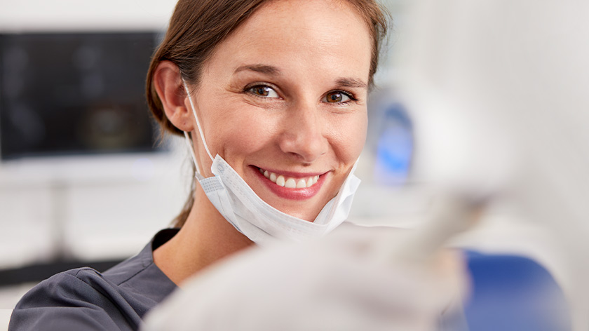 Lächelnde Mitarbeiterin der Praxis mit unter das Kinn gezogenen Mundschutz